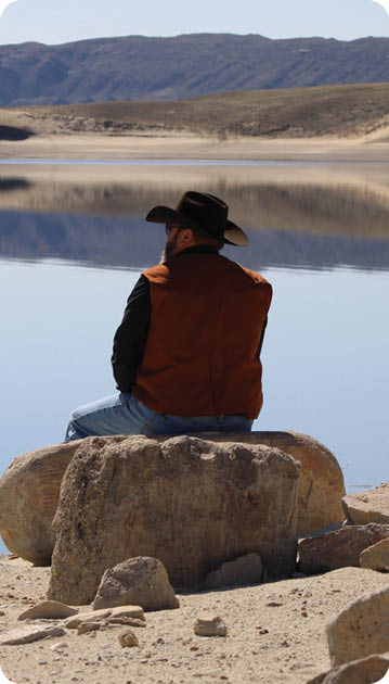 A man wearing a cowboy hat and a red vest is sitting on a rock near a body of water. AI generated content